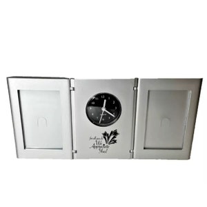 May include: Silver-colored triple picture frame with a clock in the center. The clock face is black with white numbers and hands. The frame has two picture slots on either side of the clock. The frame has an inscription that reads "For all you do, We Appreciate You!"