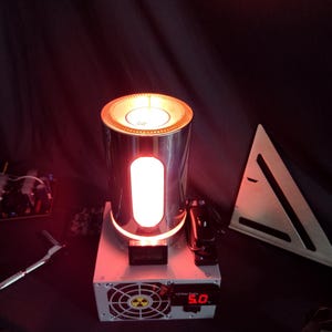 May include: A silver cylindrical lamp with a red light inside. The lamp is sitting on top of a white power supply with a black fan and a red digital display that reads "5.0".