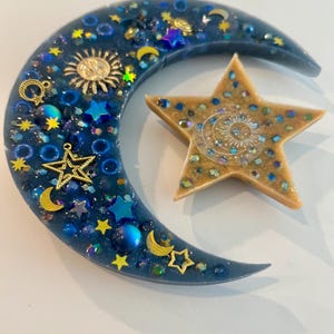 K&ouml;nnte beinhalten: Ein Kunstwerk aus Harz mit Himmelsmotiven, das einen Halbmond und einen f&uuml;nfzackigen Stern zeigt. Der Mond ist tiefblau und mit goldenen Sternen, Monden und einem Sonnenanh&auml;nger verziert. Der Stern ist beige mit eingebettetem Glitzer und einem Sonnendesign.