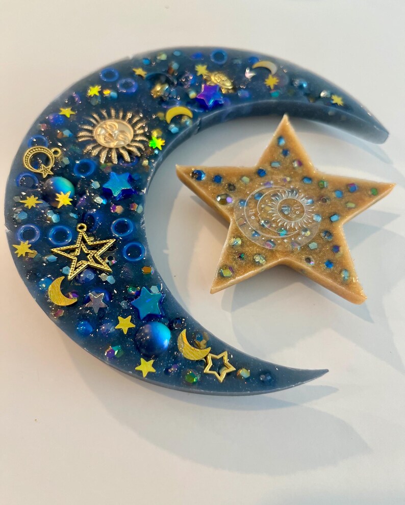 K&ouml;nnte beinhalten: Ein Kunstwerk aus Harz mit Himmelsmotiven, das einen Halbmond und einen f&uuml;nfzackigen Stern zeigt. Der Mond ist tiefblau und mit goldenen Sternen, Monden und einem Sonnenanh&auml;nger verziert. Der Stern ist beige mit eingebettetem Glitzer und einem Sonnendesign.