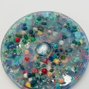 Peut inclure: Un dessous de verre rond en r&eacute;sine transparente, rempli de perles color&eacute;es, de paillettes et de petits objets bleus, verts, rouges et blancs. Un globe iris&eacute; est incrust&eacute; au centre. Un objet d&eacute;coratif.