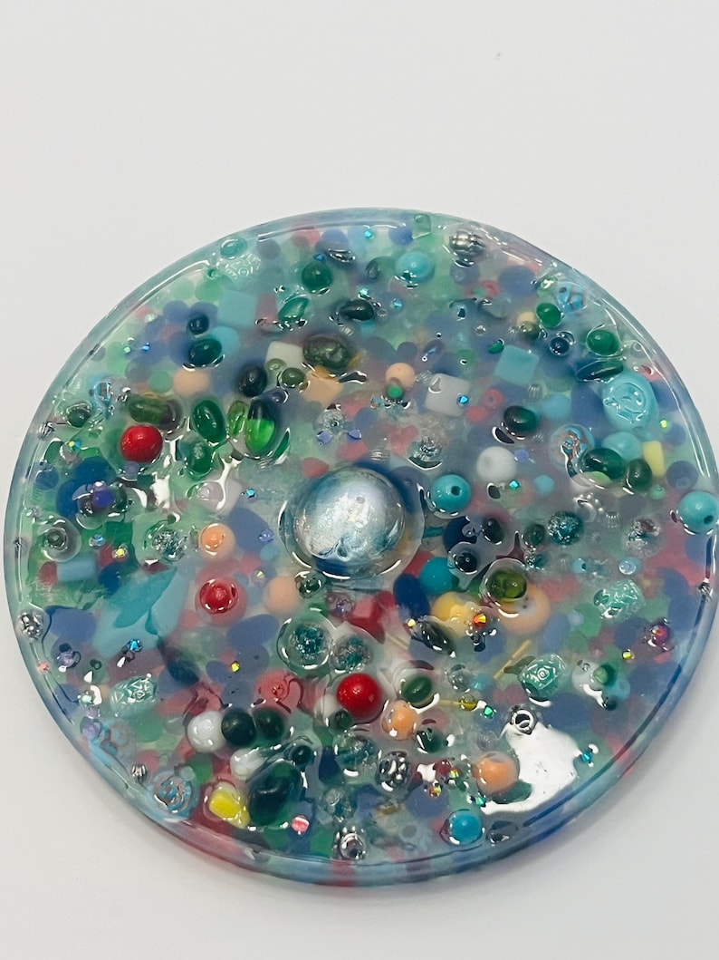 Peut inclure: Un dessous de verre rond en r&eacute;sine transparente, rempli de perles color&eacute;es, de paillettes et de petits objets bleus, verts, rouges et blancs. Un globe iris&eacute; est incrust&eacute; au centre. Un objet d&eacute;coratif.