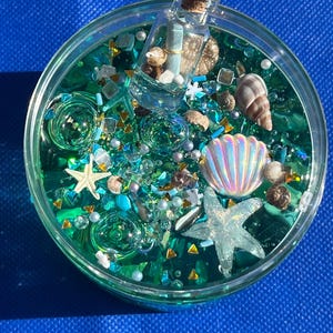 May include: A round, clear container filled with teal-colored liquid and various ocean-themed embellishments. Includes a miniature bottle with a cork, seashells, starfish, and glitter. The background is a solid blue.