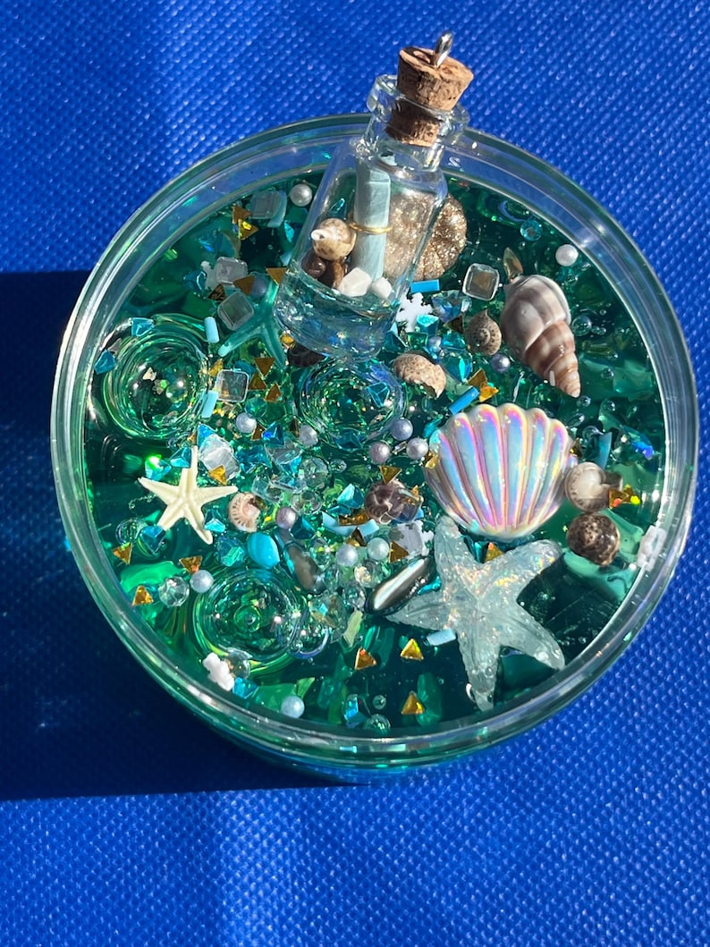 May include: A round, clear container filled with teal-colored liquid and various ocean-themed embellishments. Includes a miniature bottle with a cork, seashells, starfish, and glitter. The background is a solid blue.