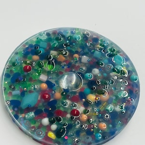 Peut inclure: Un dessous de verre rond en r&eacute;sine transparente, incrust&eacute; de nombreuses perles et &eacute;l&eacute;ments d&eacute;coratifs color&eacute;s. Le dessous de verre pr&eacute;sente des teintes bleues, vertes, rouges, oranges et blanches. Une gemme claire et ronde est incrust&eacute;e au centre.