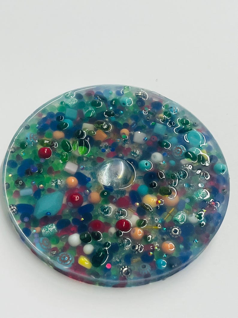 Peut inclure: Un dessous de verre rond en r&eacute;sine transparente, incrust&eacute; de nombreuses perles et &eacute;l&eacute;ments d&eacute;coratifs color&eacute;s. Le dessous de verre pr&eacute;sente des teintes bleues, vertes, rouges, oranges et blanches. Une gemme claire et ronde est incrust&eacute;e au centre.