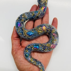 May include: A decorative snake figurine crafted from a translucent grey material, featuring an intricate surface texture embedded with small gold beads and iridescent, multi-colored crystalline accents that shimmer in shades of blue, purple, and pink.