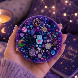 May include: A circular purple resin coaster embedded with various charms and beads, including a light blue bear with a pink heart, a crescent moon, small flowers, stars, and dark floral accents, creating a dense, multi-textured surface.