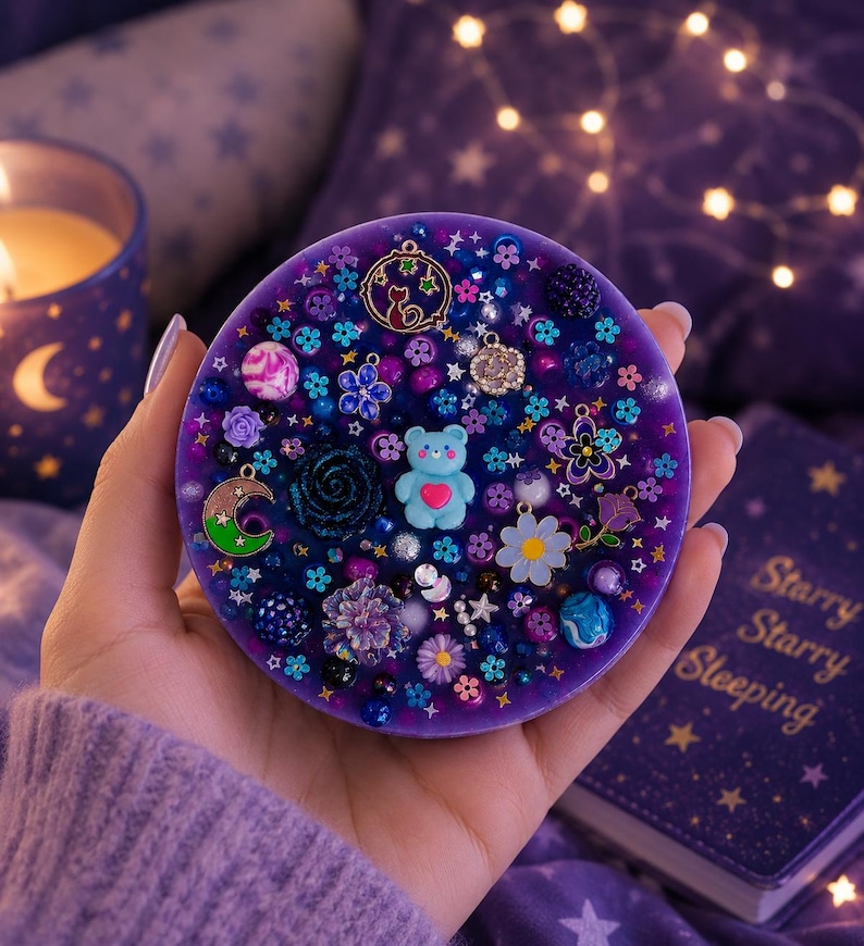 May include: A circular purple resin coaster embedded with various charms and beads, including a light blue bear with a pink heart, a crescent moon, small flowers, stars, and dark floral accents, creating a dense, multi-textured surface.