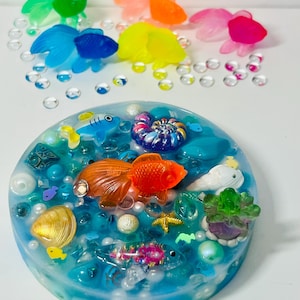 May include: A resin art piece featuring a vibrant underwater scene with an orange goldfish, seashells, and colorful fish figurines. The piece is surrounded by clear, bubble-like embellishments and additional colorful fish.