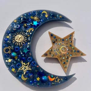 K&ouml;nnte beinhalten: Ein Kunstwerk aus Harz mit Himmelsmotiven, das einen Halbmond und einen Stern zeigt. Der dunkelblaue Mond ist mit goldenen Sonnen- und Sternanh&auml;ngern und schillernden Perlen verziert. Der Stern ist goldfarben mit einem Sonnendesign und schillernden Akzenten.
