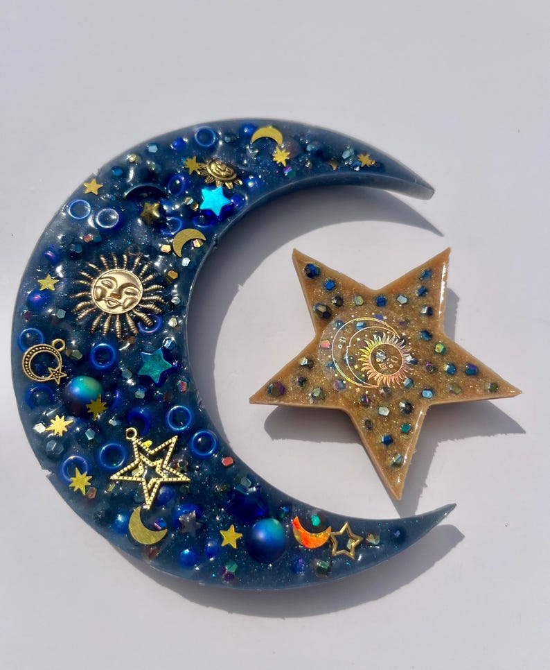 K&ouml;nnte beinhalten: Ein Kunstwerk aus Harz mit Himmelsmotiven, das einen Halbmond und einen Stern zeigt. Der dunkelblaue Mond ist mit goldenen Sonnen- und Sternanh&auml;ngern und schillernden Perlen verziert. Der Stern ist goldfarben mit einem Sonnendesign und schillernden Akzenten.
