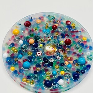 Peut inclure: Un dessous de verre rond en r&eacute;sine transparente, incrust&eacute; de perles et de bibelots color&eacute;s. Le dessous de verre pr&eacute;sente un m&eacute;lange d'&eacute;l&eacute;ments bleus, rouges, jaunes et iris&eacute;s, cr&eacute;ant une surface textur&eacute;e et d&eacute;corative. Des perles de diff&eacute;rentes tailles et formes sont int&eacute;gr&eacute;es.