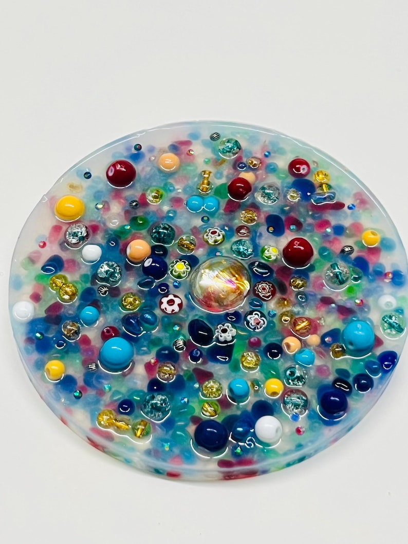 Peut inclure: Un dessous de verre rond en r&eacute;sine transparente, incrust&eacute; de perles et de bibelots color&eacute;s. Le dessous de verre pr&eacute;sente un m&eacute;lange d'&eacute;l&eacute;ments bleus, rouges, jaunes et iris&eacute;s, cr&eacute;ant une surface textur&eacute;e et d&eacute;corative. Des perles de diff&eacute;rentes tailles et formes sont int&eacute;gr&eacute;es.