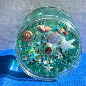 May include: A clear glass jar filled with turquoise liquid and various sea-themed decorations. The jar contains seashells, starfish, and glitter, creating a miniature ocean scene. The jar is sitting on a blue surface.