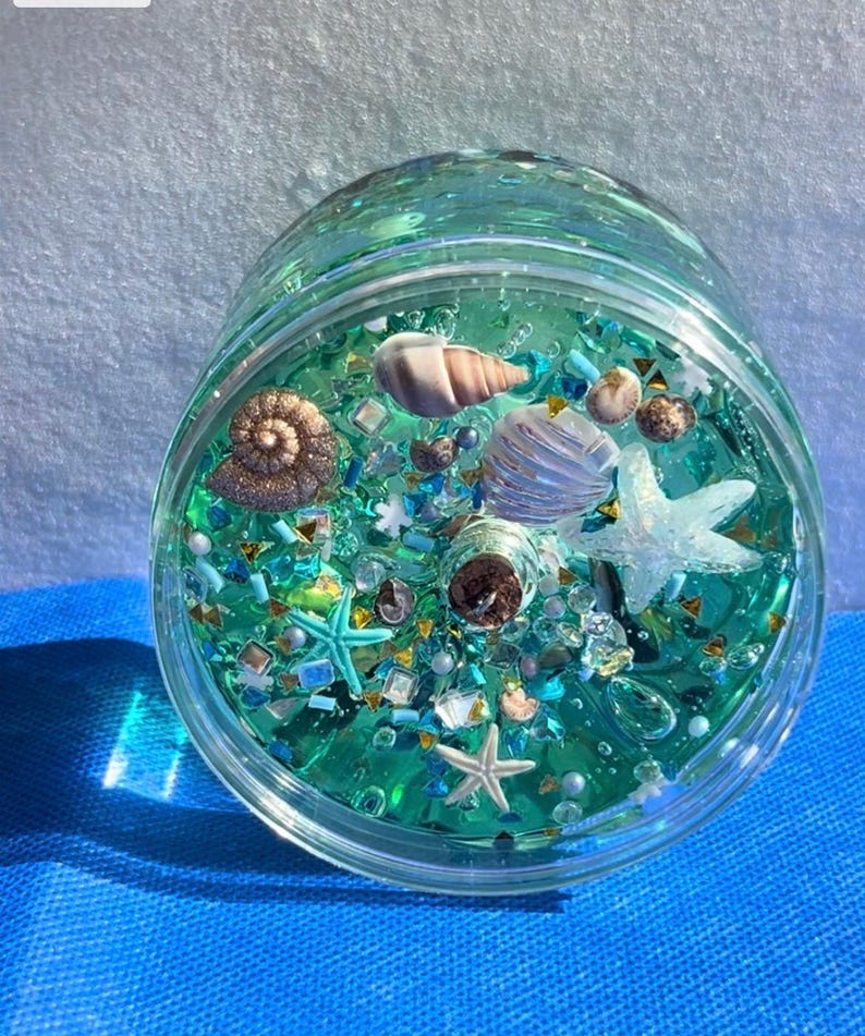 May include: A clear glass jar filled with turquoise liquid and various sea-themed decorations. The jar contains seashells, starfish, and glitter, creating a miniature ocean scene. The jar is sitting on a blue surface.