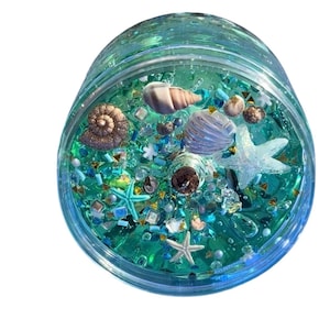 May include: A clear, round container filled with blue-green liquid and various decorative elements. The contents include seashells, starfish, and small, colorful beads. The container has a clear lid and is set against a white background.