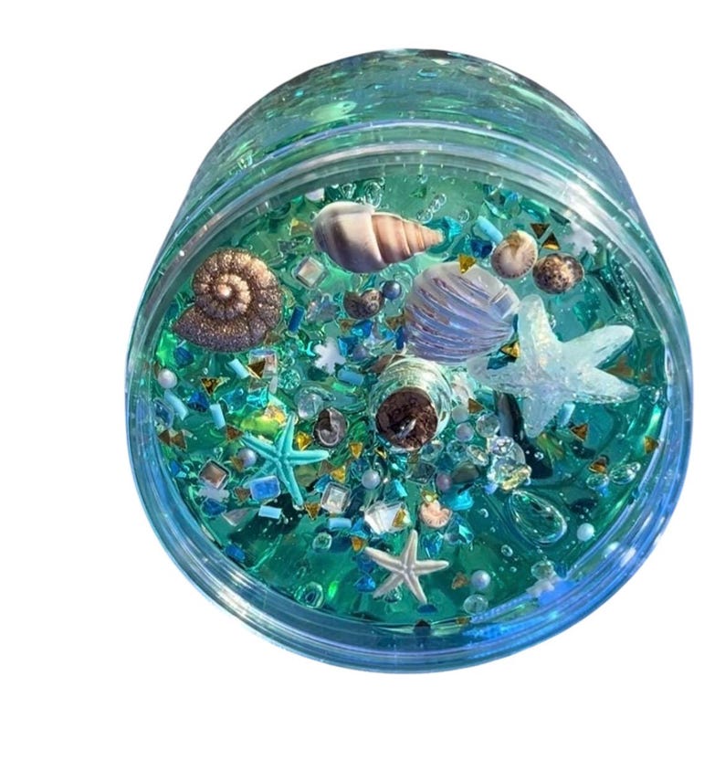 May include: A clear, round container filled with blue-green liquid and various decorative elements. The contents include seashells, starfish, and small, colorful beads. The container has a clear lid and is set against a white background.