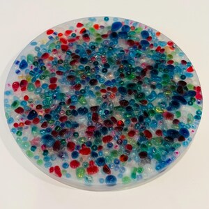 Peut inclure: Un dessous de verre rond avec une base en r&eacute;sine transparente remplie de perles color&eacute;es. Les perles sont de diff&eacute;rentes tailles et couleurs, dont le rouge, le bleu, le vert et le transparent. Le dessous de verre mesure environ 10 cm de diam&egrave;tre.