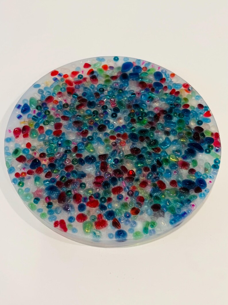 Peut inclure: Un dessous de verre rond avec une base en r&eacute;sine transparente remplie de perles color&eacute;es. Les perles sont de diff&eacute;rentes tailles et couleurs, dont le rouge, le bleu, le vert et le transparent. Le dessous de verre mesure environ 10 cm de diam&egrave;tre.