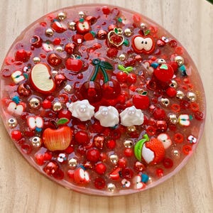 May include: A circular resin coaster featuring a translucent red base embedded with various red apple-shaped charms, small metallic gold beads, red spherical beads, and three white whipped cream-like dollops topped with small white pearls.