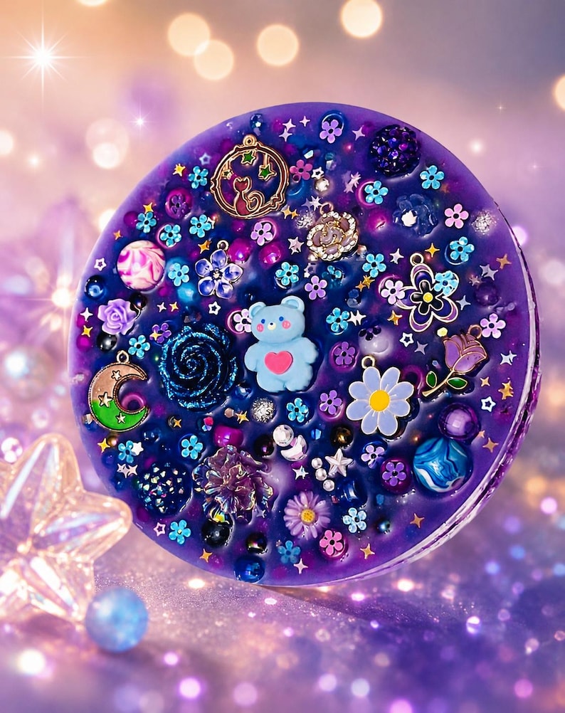 May include: A circular purple resin disc embedded with various charms including a light blue bear with a pink heart, a crescent moon, a flower with a yellow centre, a cat silhouette, and numerous small colourful beads, stars, and floral accents.