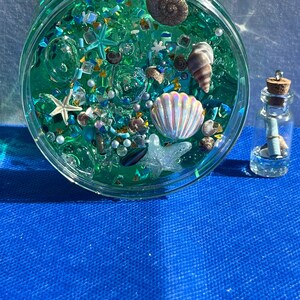 May include: A clear, round jar filled with teal-colored liquid and various decorative elements, including seashells, starfish, and iridescent beads. A small glass bottle with a cork stopper and a rolled-up paper is next to the jar. The background is a bright blue surface.