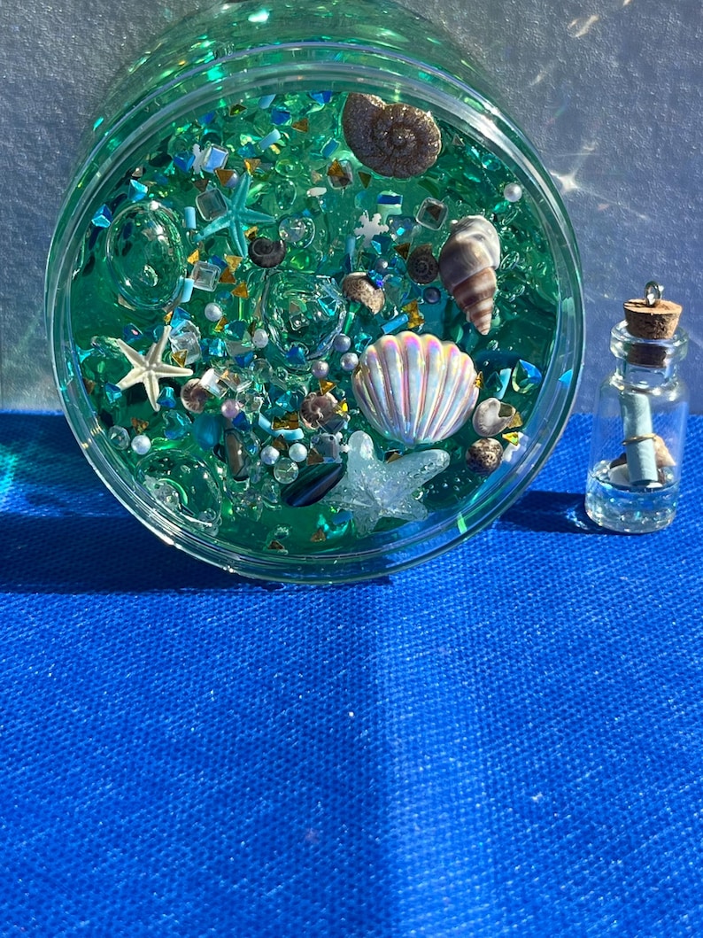 May include: A clear, round jar filled with teal-colored liquid and various decorative elements, including seashells, starfish, and iridescent beads. A small glass bottle with a cork stopper and a rolled-up paper is next to the jar. The background is a bright blue surface.