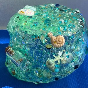 May include: A clear, rounded resin paperweight with embedded seashells, starfish, and iridescent glitter. The paperweight has a gradient of blue and green hues, with a small glass bottle attached to the side. Decorative and nautical-themed.