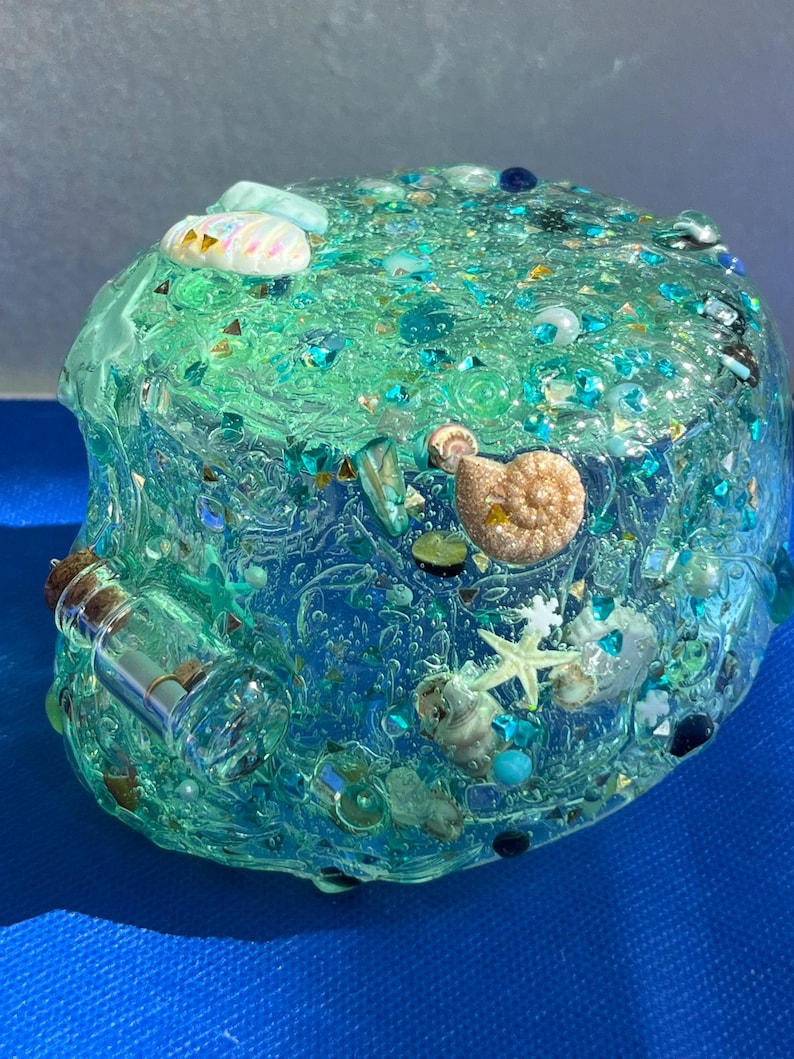 May include: A clear, rounded resin paperweight with embedded seashells, starfish, and iridescent glitter. The paperweight has a gradient of blue and green hues, with a small glass bottle attached to the side. Decorative and nautical-themed.