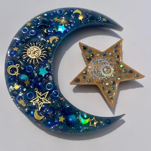 K&ouml;nnte beinhalten: Ein Kunstwerk aus Harz mit Himmelsmotiven, das einen Halbmond und einen f&uuml;nfzackigen Stern zeigt. Der Mond ist tiefblau mit eingebetteten goldenen Sternen, Monden und Sonnensymbolen. Der Stern ist goldfarben mit einem Sonnendesign und schillerndem Glitzer.