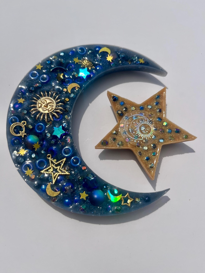 K&ouml;nnte beinhalten: Ein Kunstwerk aus Harz mit Himmelsmotiven, das einen Halbmond und einen f&uuml;nfzackigen Stern zeigt. Der Mond ist tiefblau mit eingebetteten goldenen Sternen, Monden und Sonnensymbolen. Der Stern ist goldfarben mit einem Sonnendesign und schillerndem Glitzer.