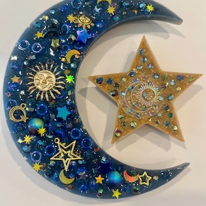 K&ouml;nnte beinhalten: Ein Kunstwerk aus Harz mit Himmelsmotiven, das einen Halbmond und einen f&uuml;nfzackigen Stern zeigt. Der Mond ist dunkelblau mit eingebetteten goldenen Sonnen- und Sternanh&auml;ngern, blauen Perlen und Glitzer. Der Stern ist goldfarben mit einem Sonnendesign.