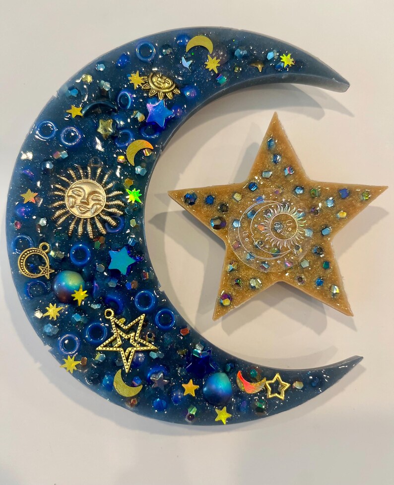 K&ouml;nnte beinhalten: Ein Kunstwerk aus Harz mit Himmelsmotiven, das einen Halbmond und einen f&uuml;nfzackigen Stern zeigt. Der Mond ist dunkelblau mit eingebetteten goldenen Sonnen- und Sternanh&auml;ngern, blauen Perlen und Glitzer. Der Stern ist goldfarben mit einem Sonnendesign.