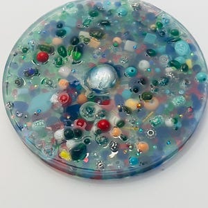 Peut inclure: Un dessous de verre rond en r&eacute;sine transparente, rempli d'une vari&eacute;t&eacute; de perles, de pierres pr&eacute;cieuses et de paillettes color&eacute;es. Le dessous de verre pr&eacute;sente un ornement sph&eacute;rique central, iris&eacute; et une multitude de petits accents color&eacute;s dans les tons de bleu, vert, rouge et orange.
