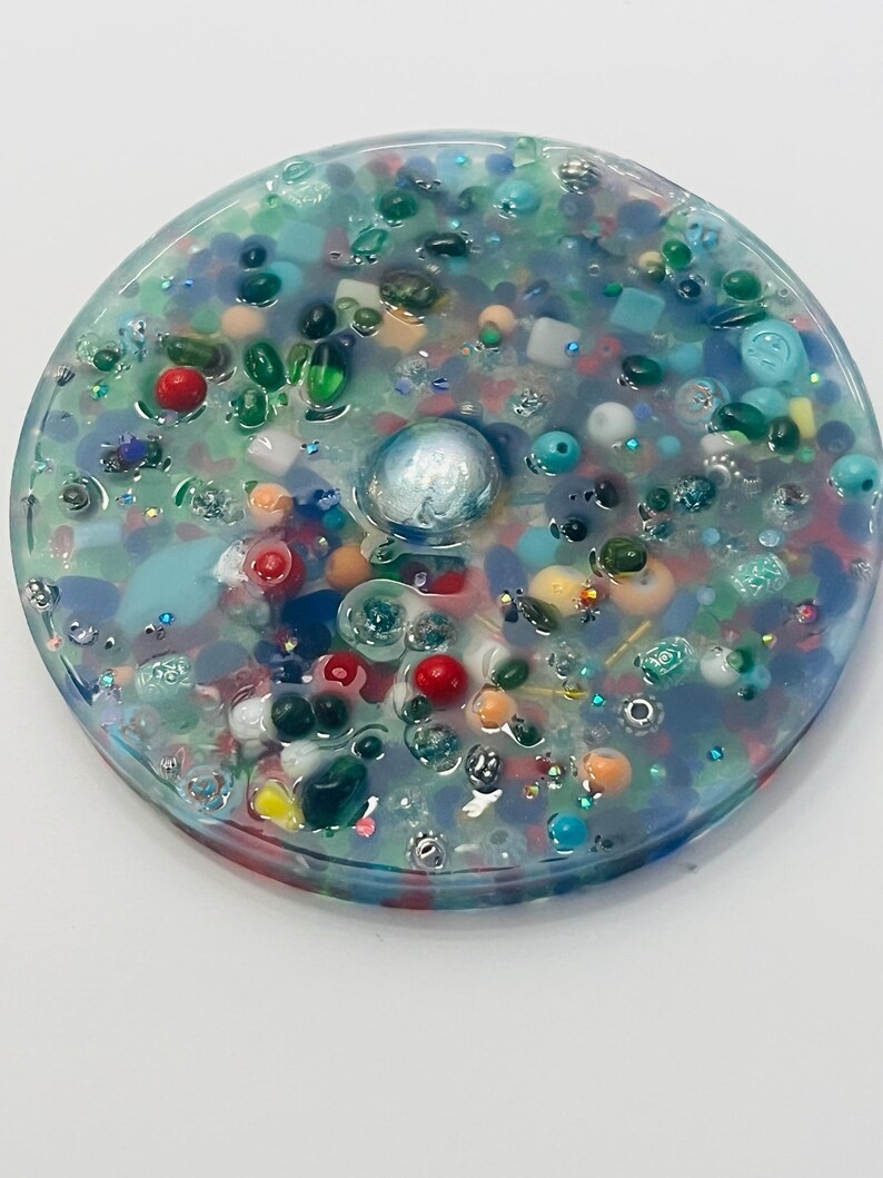 Peut inclure: Un dessous de verre rond en r&eacute;sine transparente, rempli d'une vari&eacute;t&eacute; de perles, de pierres pr&eacute;cieuses et de paillettes color&eacute;es. Le dessous de verre pr&eacute;sente un ornement sph&eacute;rique central, iris&eacute; et une multitude de petits accents color&eacute;s dans les tons de bleu, vert, rouge et orange.