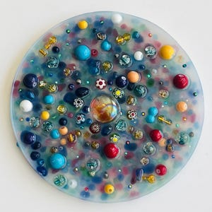 Peut inclure: Une &oelig;uvre d'art en r&eacute;sine circulaire, orn&eacute;e d'une multitude de perles et d'ornements color&eacute;s. L'&oelig;uvre pr&eacute;sente un m&eacute;lange de perles bleues, rouges, jaunes et transparentes, ainsi que de petits motifs floraux. Le design est une mosa&iuml;que vibrante et textur&eacute;e, d'environ 10 cm de diam&egrave;tre.