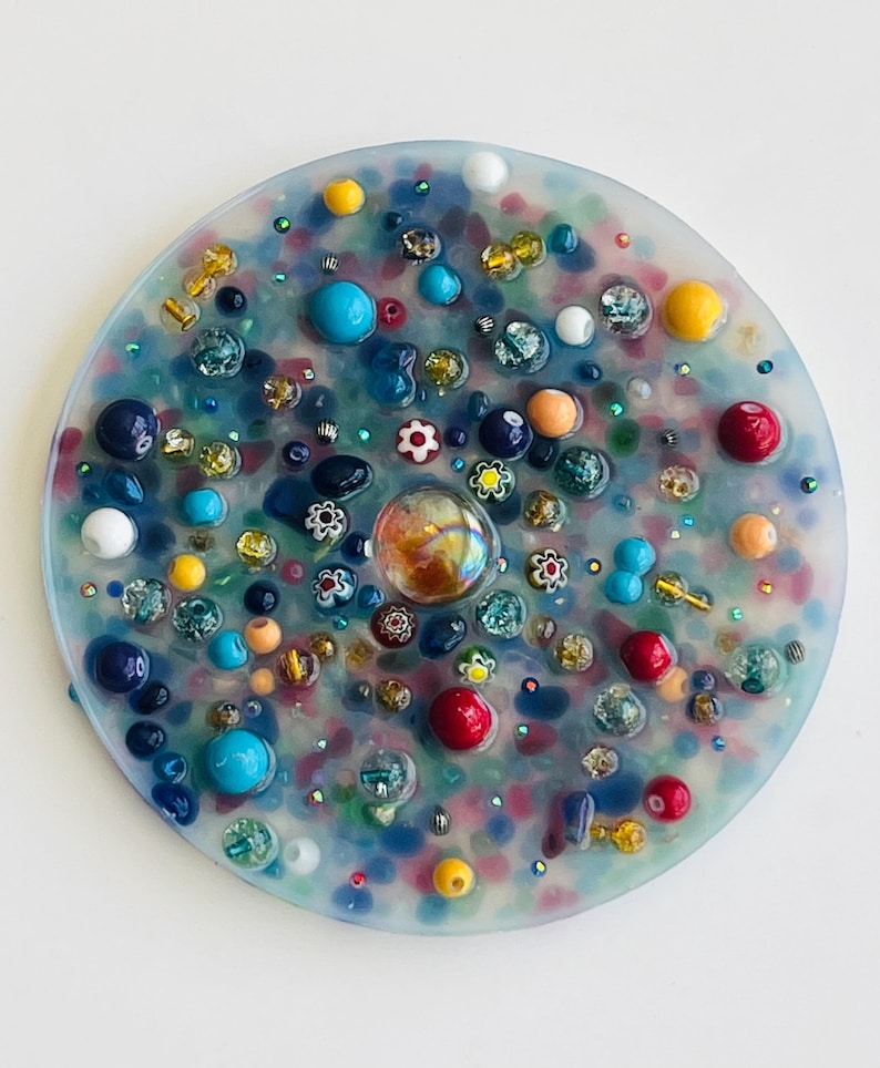 Peut inclure: Une &oelig;uvre d'art en r&eacute;sine circulaire, orn&eacute;e d'une multitude de perles et d'ornements color&eacute;s. L'&oelig;uvre pr&eacute;sente un m&eacute;lange de perles bleues, rouges, jaunes et transparentes, ainsi que de petits motifs floraux. Le design est une mosa&iuml;que vibrante et textur&eacute;e, d'environ 10 cm de diam&egrave;tre.