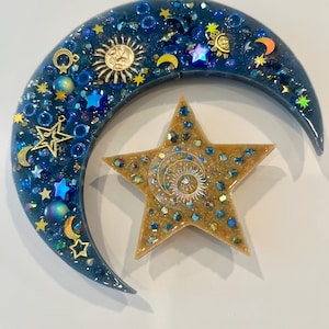 K&ouml;nnte beinhalten: Ein Kunstwerk aus Harz mit Himmelsmotiven, das einen Halbmond und einen f&uuml;nfzackigen Stern zeigt. Der dunkelblaue Mond ist mit goldenen Sonnen- und Sternanh&auml;ngern, blauen Perlen und schillernden Akzenten verziert. Der Stern ist goldfarben mit einem Sonnendesign in der Mitte.