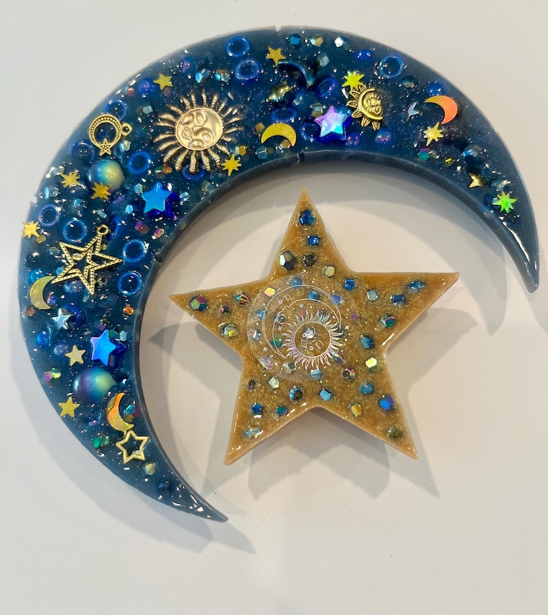 K&ouml;nnte beinhalten: Ein Kunstwerk aus Harz mit Himmelsmotiven, das einen Halbmond und einen f&uuml;nfzackigen Stern zeigt. Der dunkelblaue Mond ist mit goldenen Sonnen- und Sternanh&auml;ngern, blauen Perlen und schillernden Akzenten verziert. Der Stern ist goldfarben mit einem Sonnendesign in der Mitte.