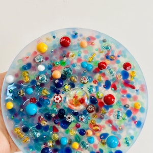 Peut inclure: Un dessous de verre rond en r&eacute;sine transparente, orn&eacute; de perles et d'&eacute;l&eacute;ments d&eacute;coratifs color&eacute;s. Il pr&eacute;sente des perles rouges, bleues, jaunes et blanches, ainsi que de petits motifs floraux. Le fond est un m&eacute;lange de couleurs.