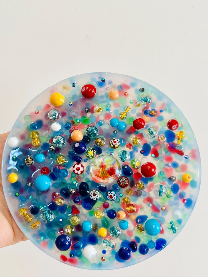 Peut inclure: Un dessous de verre rond en r&eacute;sine transparente, orn&eacute; de perles et d'&eacute;l&eacute;ments d&eacute;coratifs color&eacute;s. Il pr&eacute;sente des perles rouges, bleues, jaunes et blanches, ainsi que de petits motifs floraux. Le fond est un m&eacute;lange de couleurs.