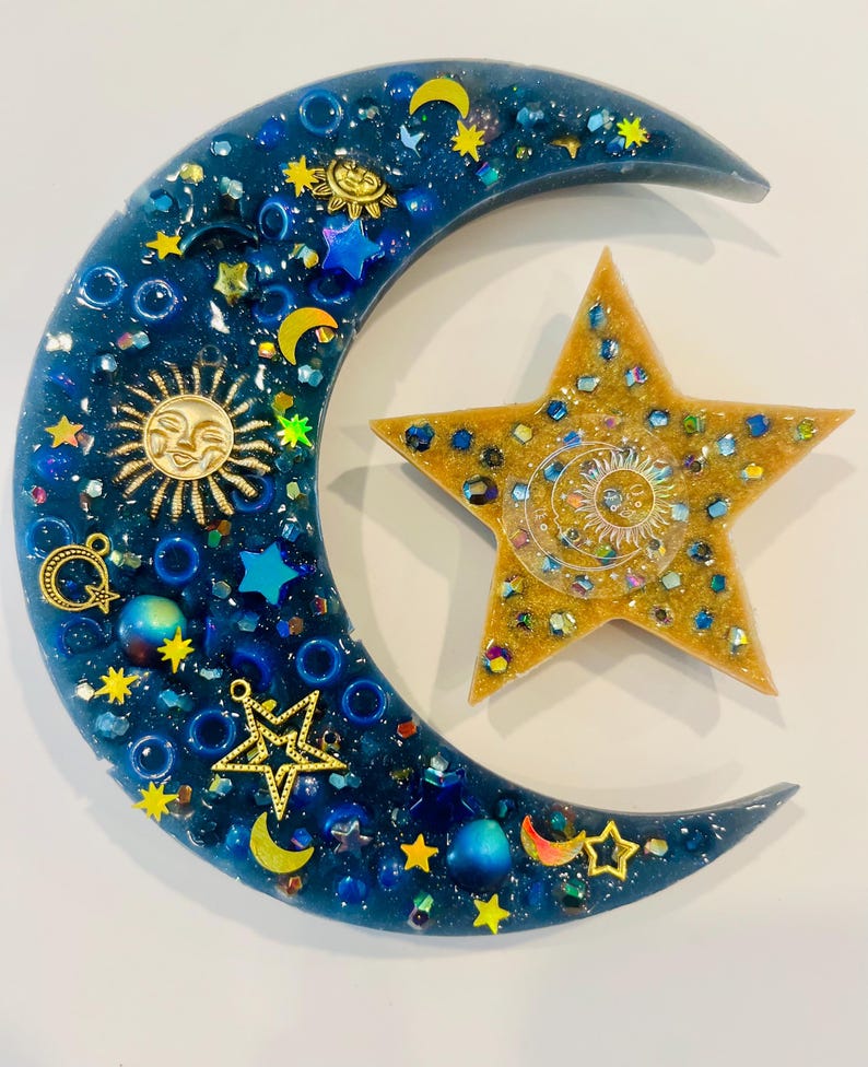 K&ouml;nnte beinhalten: Ein Kunstwerk aus Harz mit Himmelsmotiven, das einen Halbmond und einen f&uuml;nfzackigen Stern zeigt. Der Mond ist tiefblau, mit goldenen Sonnen- und Sternanh&auml;ngern verziert und mit blauem und goldenem Glitzer versehen. Der Stern ist goldfarben mit einem Sonnendesign.