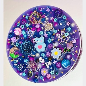 May include: A circular purple resin piece embedded with various charms including a light blue bear with a pink heart, a crescent moon, a rose, flowers, stars, and assorted beads. The surface is filled with colourful decorative elements.