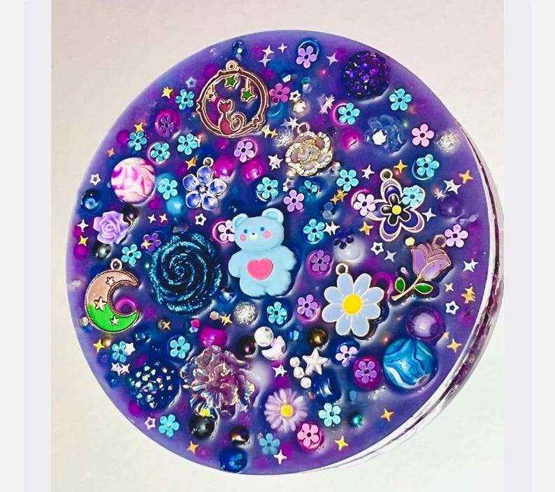 May include: A circular purple resin piece embedded with various charms including a light blue bear with a pink heart, a crescent moon, a rose, flowers, stars, and assorted beads. The surface is filled with colourful decorative elements.