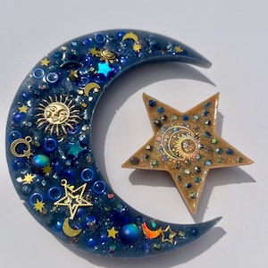 K&ouml;nnte beinhalten: Ein Kunstwerk aus Harz mit Himmelsmotiven, das einen Halbmond und einen Stern zeigt. Der dunkelblaue Mond ist mit goldenen Sonnen- und Sternanh&auml;ngern und schillernden Perlen verziert. Der Stern ist goldfarben mit einem Sonnen- und Monddesign.