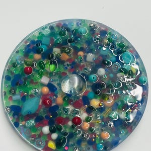 May include: A round, clear resin coaster filled with colorful beads and small objects. The coaster features a variety of colors, including red, green, blue, and yellow, with a clear, round gem in the center. The design gives the appearance of a miniature underwater scene.