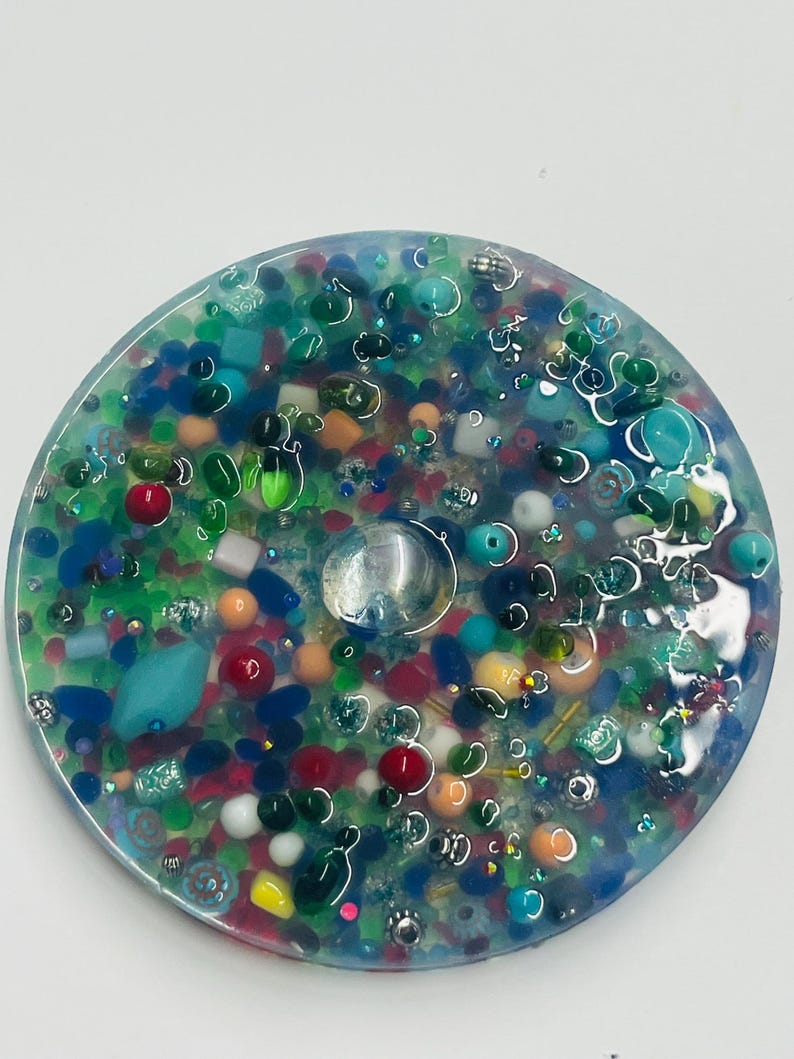 Peut inclure: Un dessous de verre rond en r&eacute;sine transparente, rempli de perles et de petits objets color&eacute;s. Le dessous de verre pr&eacute;sente une vari&eacute;t&eacute; de couleurs, notamment le rouge, le vert, le bleu et le jaune, avec un bijou rond et transparent au centre. Le design &eacute;voque une sc&egrave;ne sous-marine miniature.