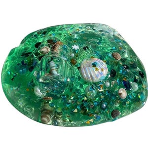 May include: A vibrant green slime with embedded miniature seashells, iridescent glitter, and a small glass bottle containing a miniature ship. The slime is a translucent green color, with various shades of blue and green glitter.