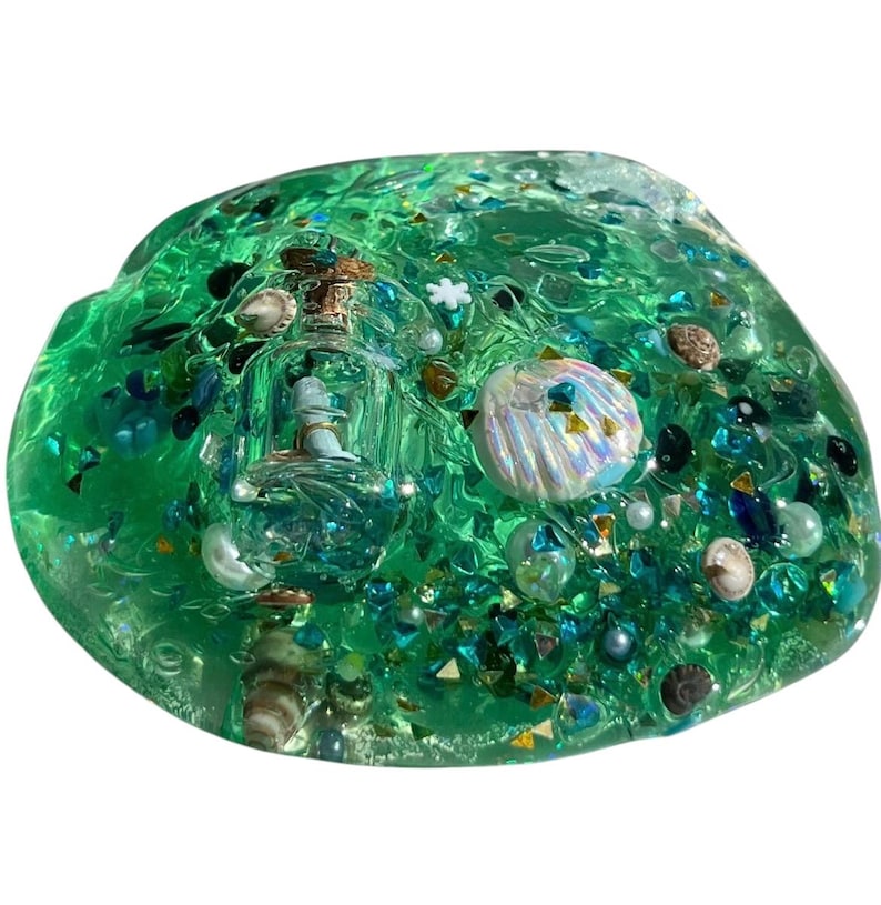 May include: A vibrant green slime with embedded miniature seashells, iridescent glitter, and a small glass bottle containing a miniature ship. The slime is a translucent green color, with various shades of blue and green glitter.